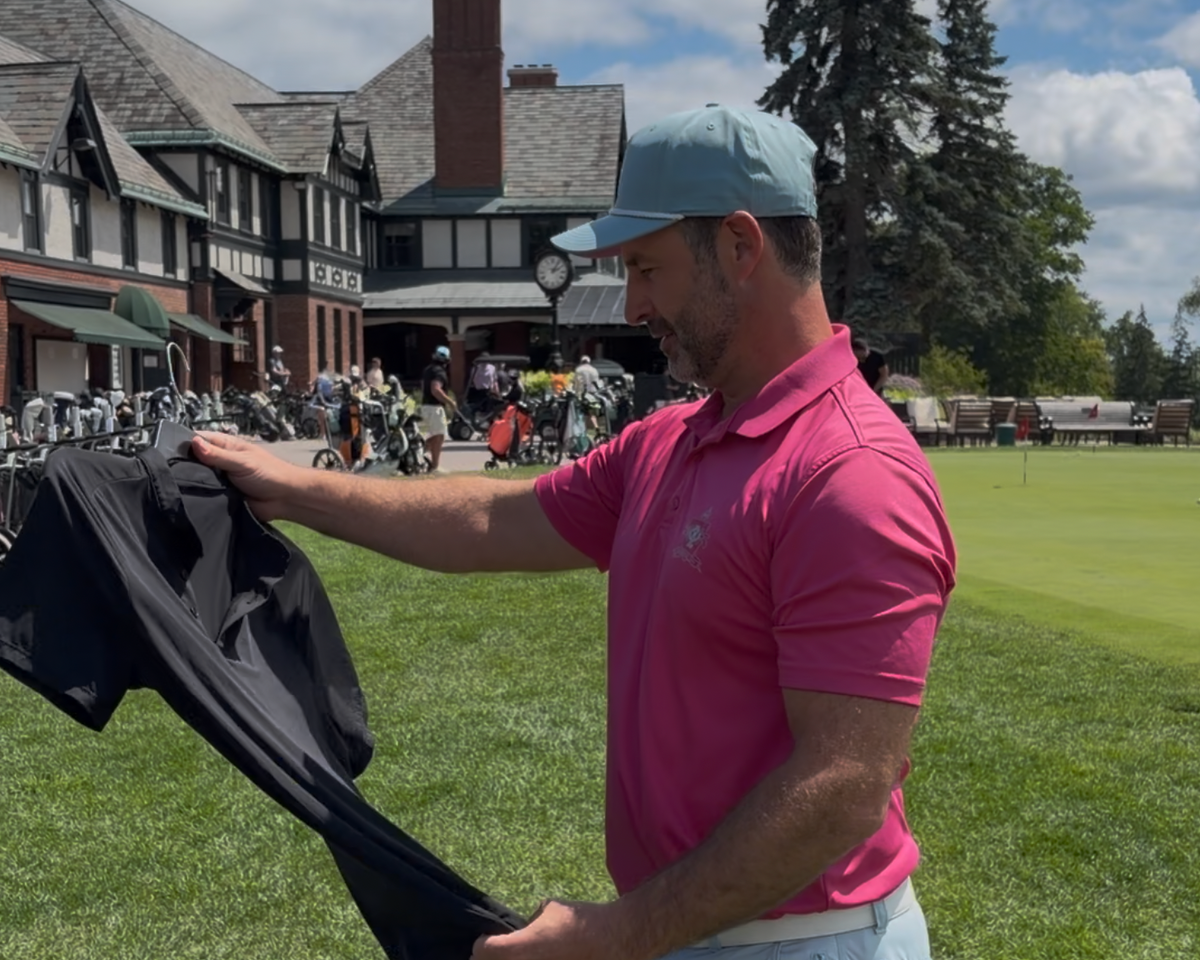 A man in a pink polo shirt and blue cap holds up a black golf shirt on a hanger in a grassy outdoor setting with a clubhouse in the background.
